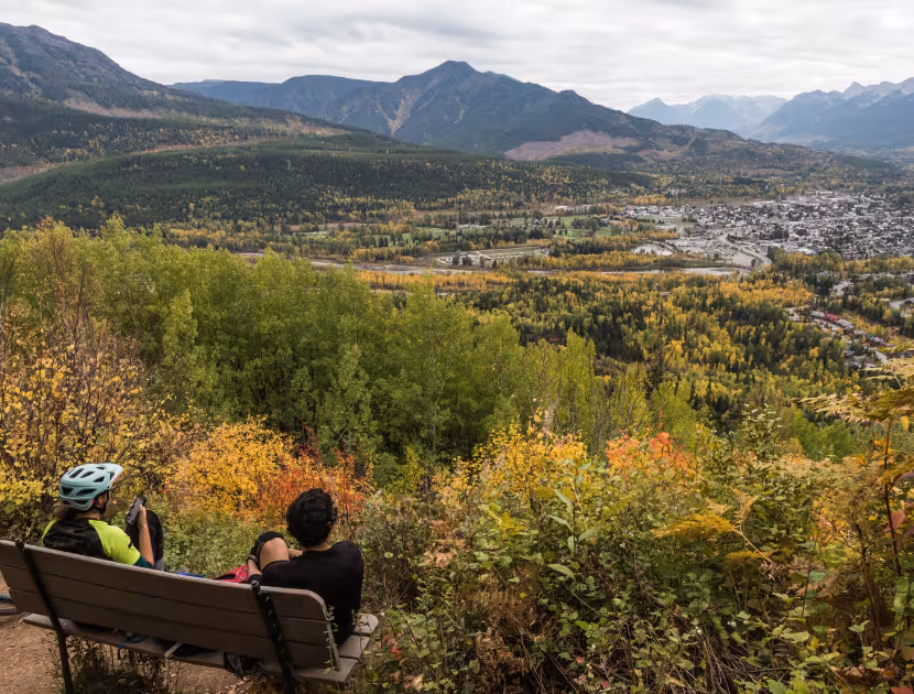 Two people sitting on a bench overlooking a sunlit mountain valley town.