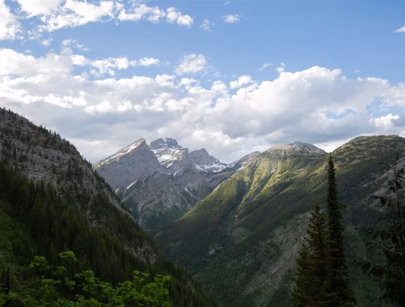 Mountainous landscape with lush greenery, snow-capped peaks of The Three Sisters mountain in Fernie BC, and blue sky with white clouds.