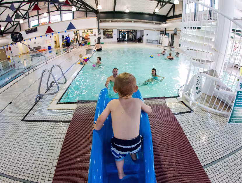 Boy climbing blue waterslide toward indoor pool with people swimming.