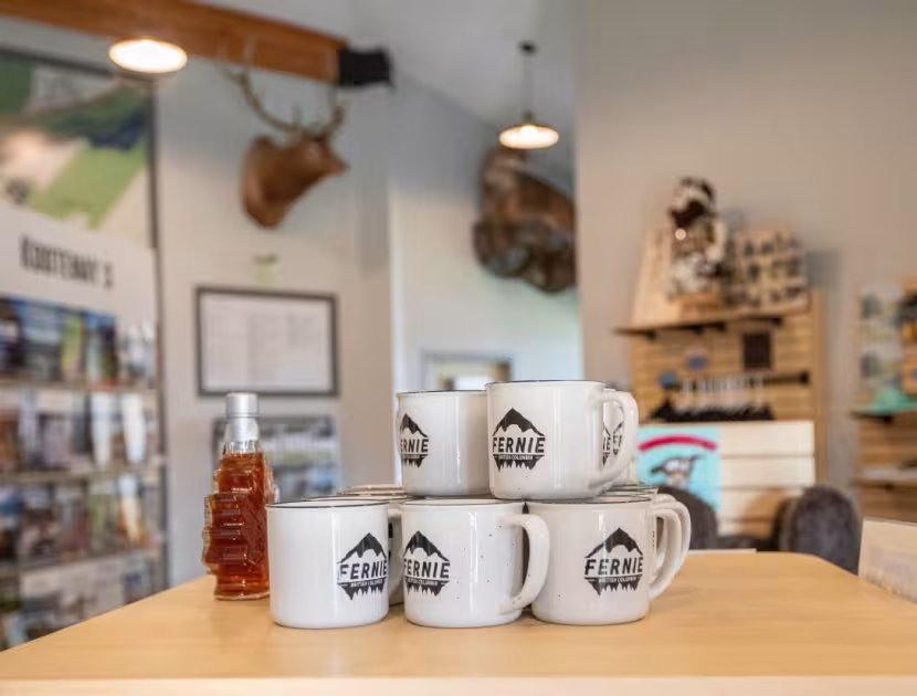 Stack of white mugs with a logo on a wooden table, bottle beside them; blurred store background.