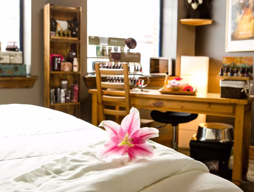 A blurred view of a bed with a white comforter and pink flower on top, with a wooden desk and chair in the background.