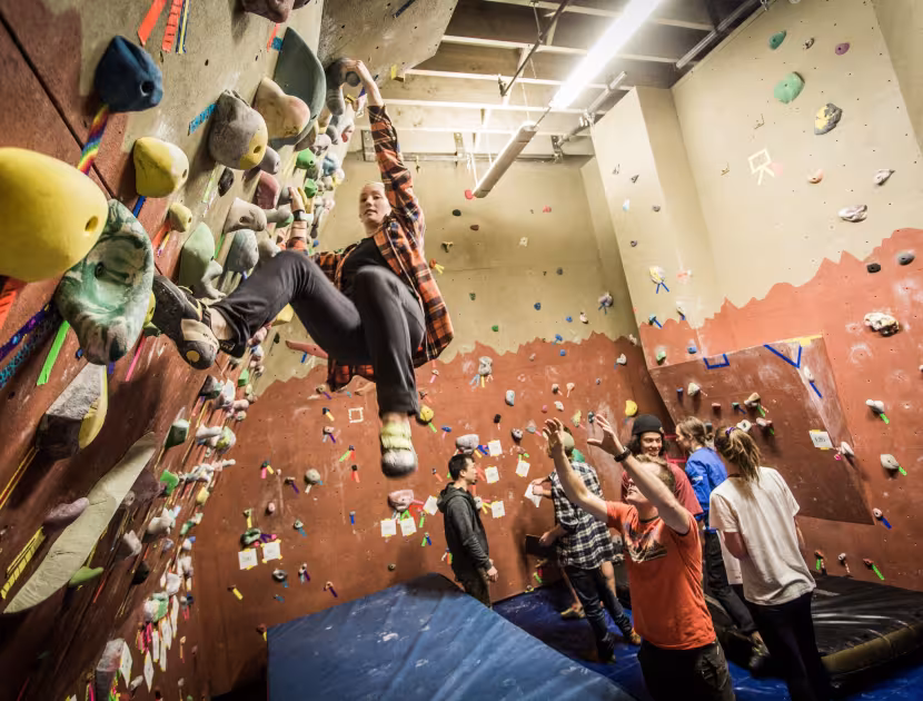 Indoor rock climbing wall with people climbing and watching; colorful holds, beige and reddish-brown walls, blue floor mats.