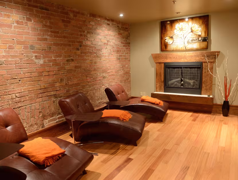 Cozy relaxation room with 3 brown leather recliners, orange pillows, brick wall, fireplace, and wooden floor.