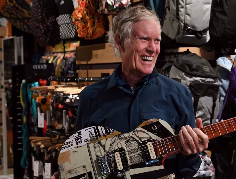 Man playing a camouflage-patterned guitar in a store with outdoor gear.