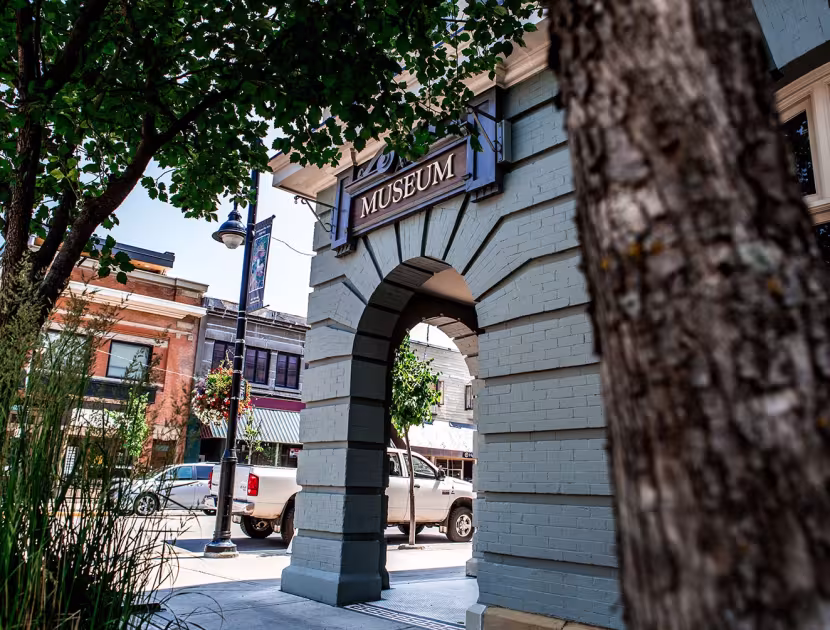 Stone archway with a sign. Tree trunk and sidewalk; background buildings.