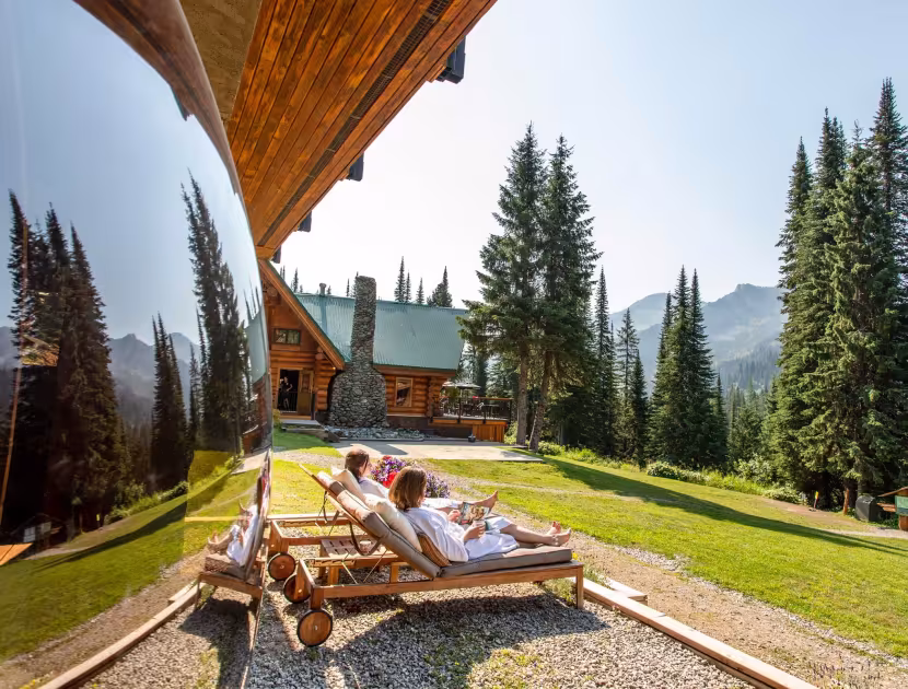 Two people in bathrobes relaxing on wooden lounge chairs under a wooden awning, with a large lodge and evergreen trees in the background.