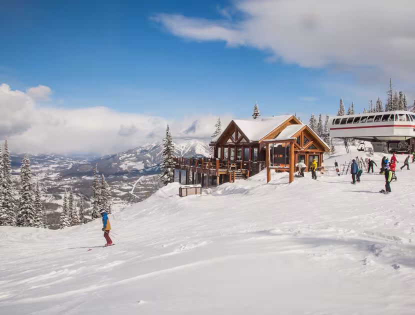 Ski resort with snow-covered log building, trees, and mountains under a blue cloudy sky.