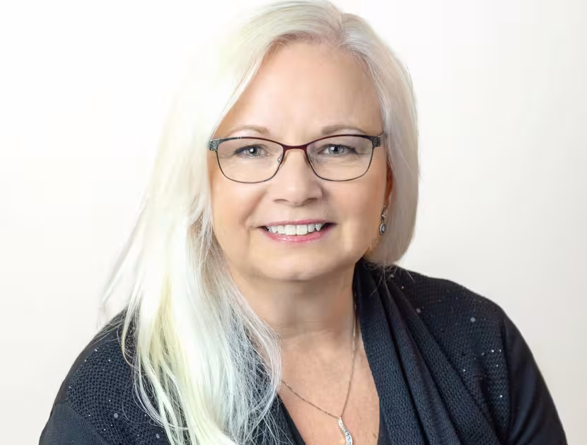 Headshot of a woman with long white hair, wearing glasses and a black top, smiling.