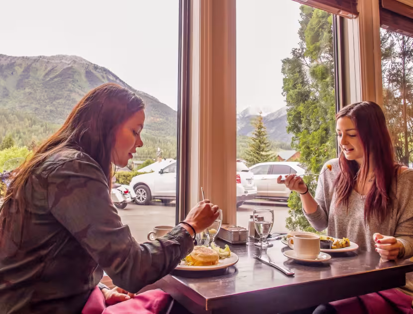 Two women eating breakfast at a table by a large window with a mountain view.