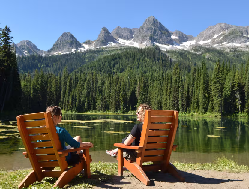 Two people sitting on wooden chairs overlooking a serene alpine lake and snow-capped mountains.