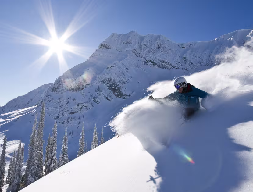 Skier in blue jacket speeding down snowy slope with mountain in background.