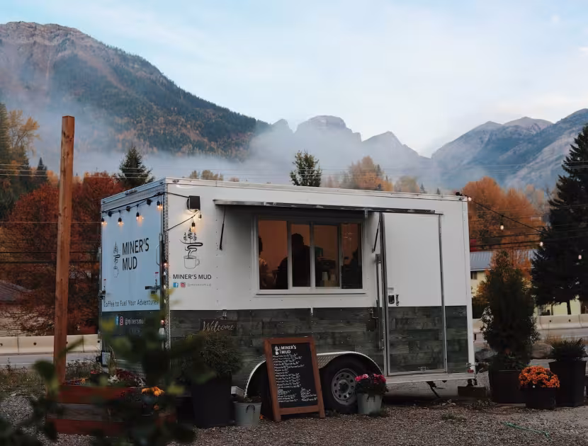 White food truck parked in front of mountains, surrounded by plants and trees.