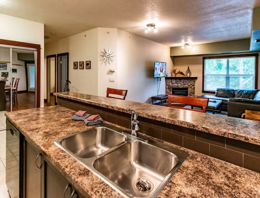 Kitchen with brown marbled countertop and stainless steel sink, adjacent to living room with brown couch and TV above fireplace.