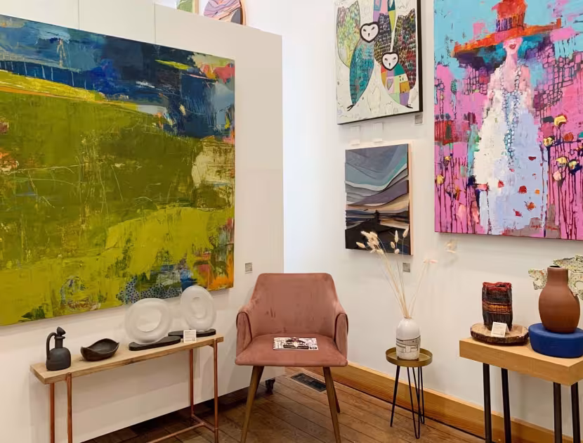 A cozy art gallery interior with paintings on a white wall; a pink chair, and decorative items on wooden tables.