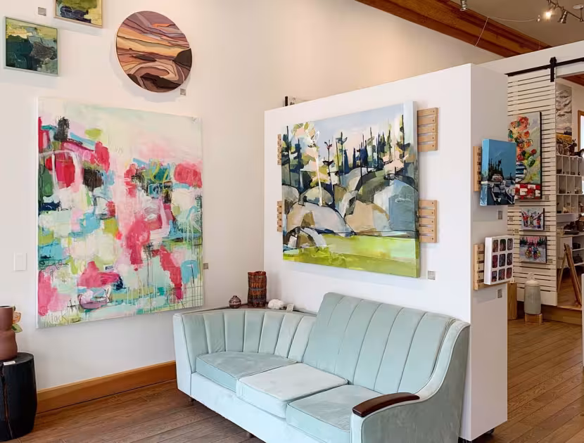 Cozy art gallery interior with light blue sofa, wooden floors, and vibrant paintings on white walls.